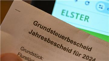 Ein Grundsteuerbescheid für 2024 wird vor einen Computerbildschirm gehalten, auf dem das Logo der Steuerplattform Elster angezeigt wird.