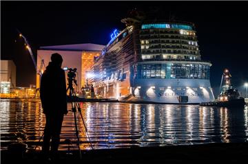 Ein Mann mit einer Filmkamera auf einem Stativ steht vor dem Hafenbecken und beobachtet das Kreuzfahrtschiff "Spectrum of the Seas" 