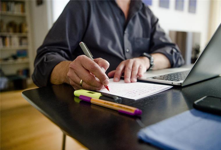 Ein Mann sitzt zuhause an einem Esstisch. Er schreibt mit einem Stift auf ein Blatt Papier neben einem Laptop.
