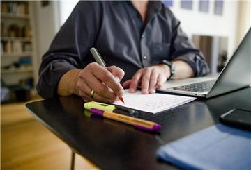 Ein Mann sitzt zuhause an einem Esstisch. Er schreibt mit einem Stift auf ein Blatt Papier neben einem Laptop.