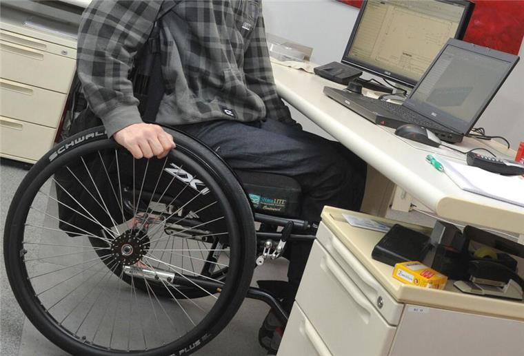 Ein Mann sitzt in einem Rollstuhl an seinem Arbeitsplatz am Schreibtisch.