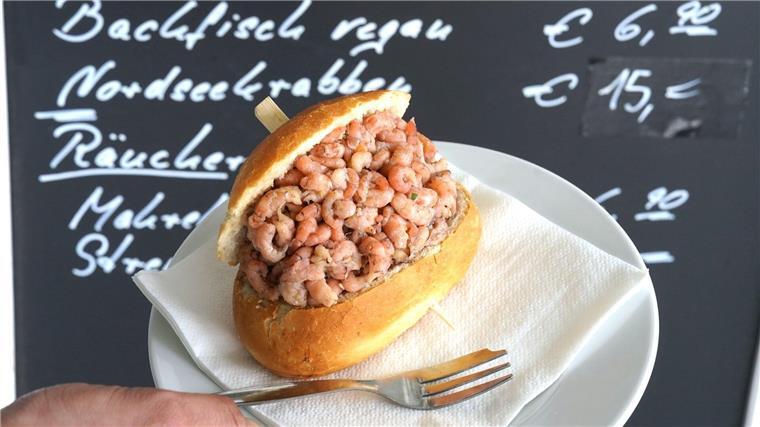 Wird das Krabbenbrötchen zum Luxusgut? Ein Mitarbeiter hält ein Nordseekrabben-Brötchen vor einer Preistafel „Nordseekrabben - 15,- Euro“.