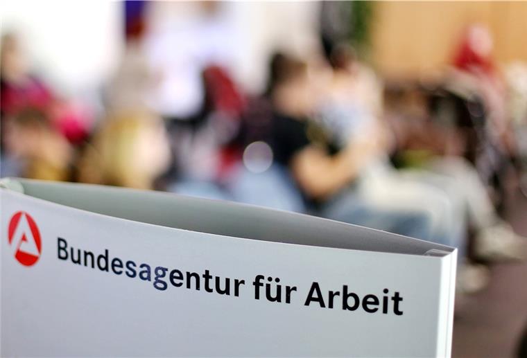 Ein Schild mit der Aufschrift „Bundesagentur für Arbeit" steht einem vollen Wartezimmer im Jobcenter.