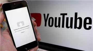 Ein Upload-Fenster der Video-Plattform Youtube ist auf einem Smartphone vor einem YouTube-Logo auf einem Bildschirm zu sehen. 