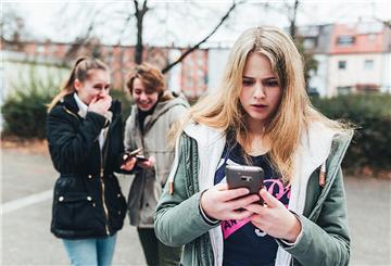  Ein Mädchen steht im Hof einer Schule und schaut auf ihr Smartphone, während im Hintergrund zwei andere Mädchen lachen 