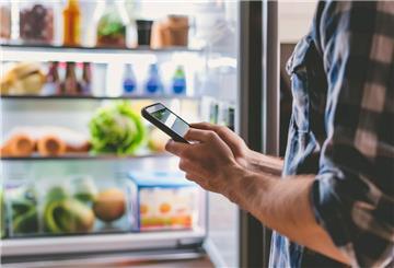 Mann steht mit Handy vor dem offenen Kühlschrank.