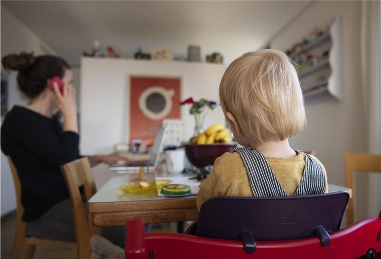 Eine Frau sitzt im Homeoffice an ihrem Laptop und telefoniert, während ihr Kind neben ihr in einem Kinderstuhl am Tisch sitzt – eine Situation, die Eltern ihren Kindern erklären sollten.