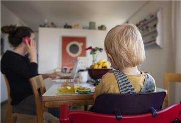 Eine Frau sitzt im Homeoffice an ihrem Laptop und telefoniert, während ihr Kind neben ihr in einem Kinderstuhl am Tisch sitzt – eine Situation, die Eltern ihren Kindern erklären sollten.
