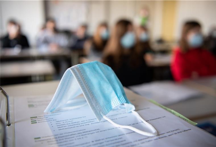 Maskenpflicht ab Donnerstag ab Klasse 5 in Bremerhaven Eine Maske liegt im Unterricht auf Unterlagen, während im Hintergrund Schülerinnen und Schüler mit Mund- und Nasenschutz dem Unterricht folgen. Foto: Matthias Balk/dpa