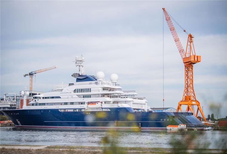 Eine riesige Yacht liegt im Hafen. Im Hintergrund sind Kräne zu sehen.