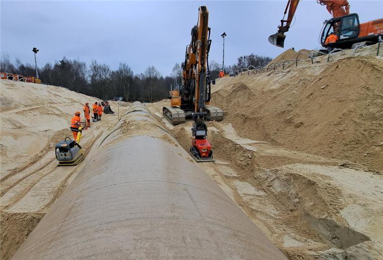 Eine Rüttelplatte wird von einem Bauarbeiter gezogen, die andere von einem Bagger: Schicht für Schicht wird der Sand um das neue Rohr verdichtet.
