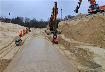 Eine Rüttelplatte wird von einem Bauarbeiter gezogen, die andere von einem Bagger: Schicht für Schicht wird der Sand um das neue Rohr verdichtet.
