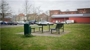 Im Vordergrund Rasen sowie zwei Bänke und ein Tisch, im Hintergrund ein mit Pkw belegter Parkplatz