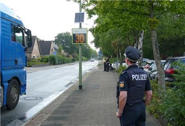 Ein Lkw fährt auf eine Geschwindigkeitsanzeige zu, 30 km/h wird angezeigt, Polizisten schauen zu.