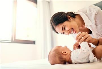 Eine dunkelhaarige Frau lacht ein auf dem Rücken liegendes Baby an.