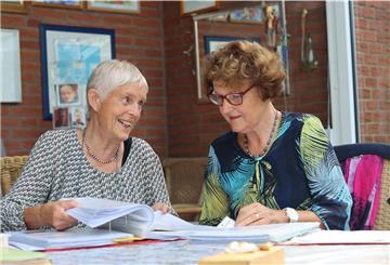 Zwei Frauen sitzen vor einem Stapel Papier und sprechen miteinander.