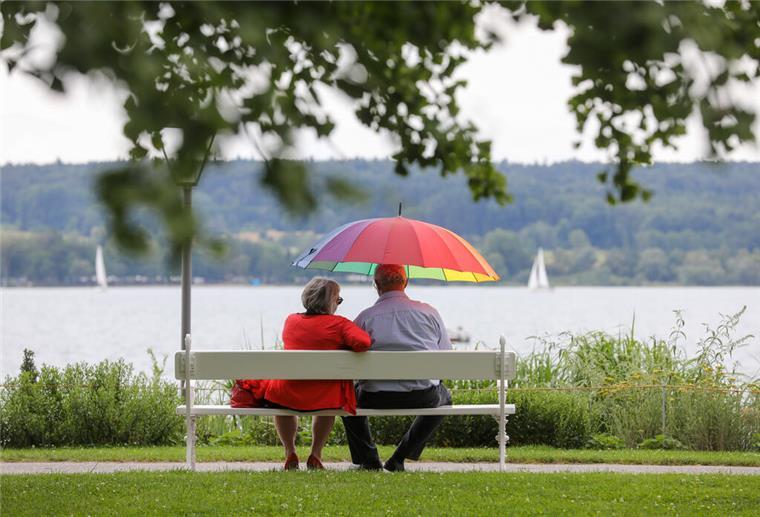 Zwei Rentner sitzen auf einer Parkbank mit Blick auf einen See. Einer von ihnen hält einen bunten Schirm.