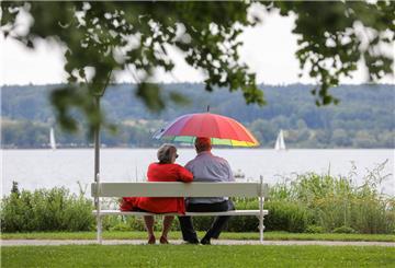 Zwei Rentner sitzen auf einer Parkbank mit Blick auf einen See. Einer von ihnen hält einen bunten Schirm.