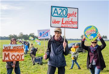 Die Demonstranten Claudia (l-r), Wolfgang und Monika,stehen mit Schildern vor einer Menschenmenge im Moor, auf denen (l-r) „we want Moor“, KüstenautoWahn“, „A20 geht über Leichen“ und „Klima retten“ zu sehen ist.