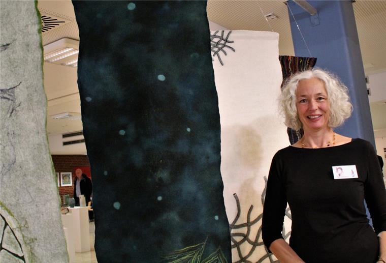 Eine Frau mit grauen Haaren steht vor einem Kunstwerk aus Filz.