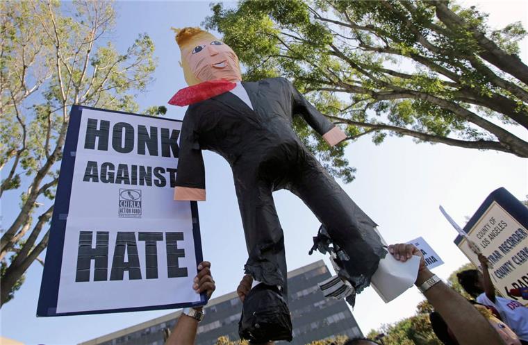 Gegner von Donald Trump halten ein Schild mit der Aufforderung "Hupe gegen Hass" und eine aufblasbare Puppe, die Donald Trump darstellt, in die Höhe. 