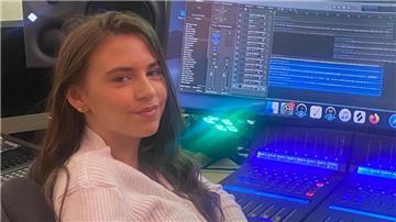 Eine junge Frau sitzt an einem Mischpult vor einem Monitor und schaut in die Kamera. 