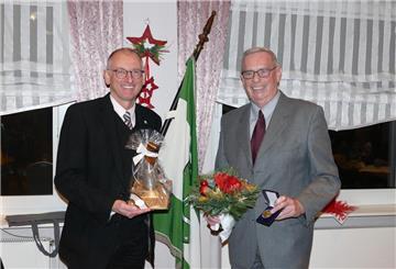 Einem Mann werden von einem anderen Mann ein Blumenstrauß, ein Ehrenbecher und eine Medaille überreicht.