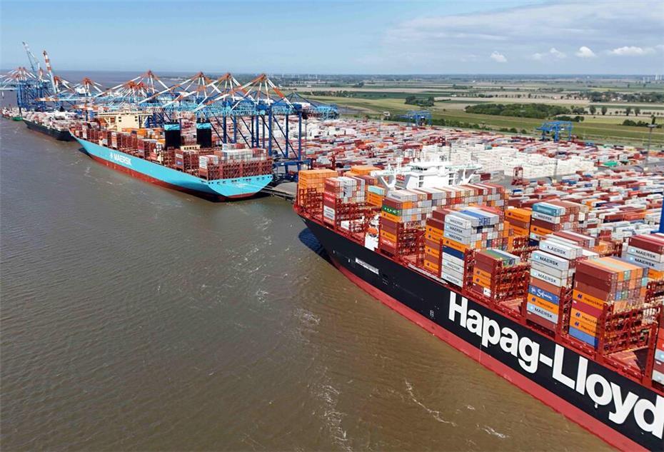 Zwei große Containerschiffe der Reedereien Maersk und Hapag-Lloyd liegen nebeneinander am Kai eines Containerterminals. Im Hintergrund sind zahlreiche Kräne und Containerstapel sowie eine weite, flache Küstenlandschaft zu sehen.
