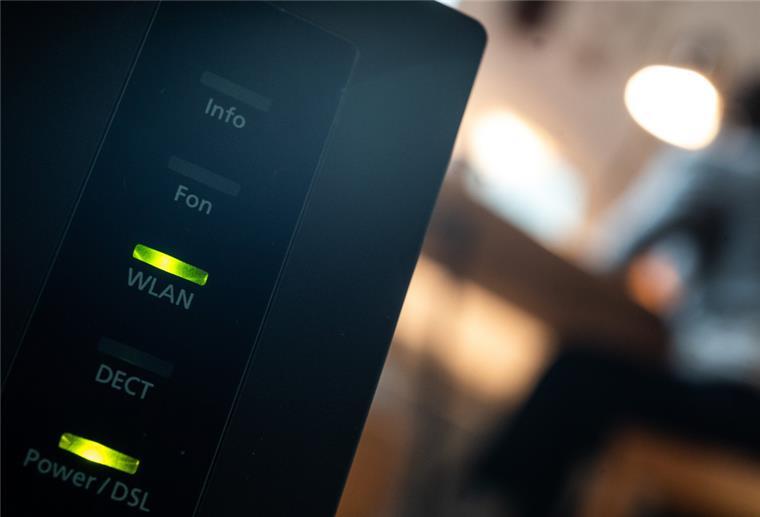 Ein Router, auf dem die LED für W-Lan und DSL leuchten, ist in einer Wohung zu sehen, in der eine Person an einem Tisch sitzt.