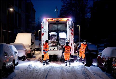 Mitarbeiter der Abfallwirtschaft leeren am frühen Morgen die Mülltonnen in einer völlig verschneiten Straße