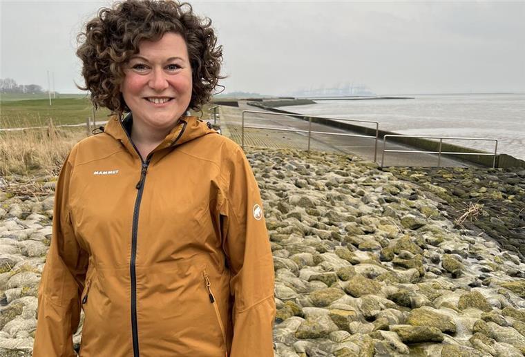 Frau mit braunen Locken steht an der Küste, im Hintergrund steht ein kleiner schwarz-weiß gestreifter Leuchtturm.