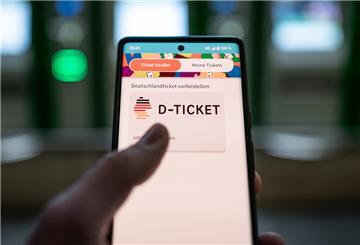 Ein Mann hält ein Mobiltelefon, auf dem das „D-Ticket“ gekauft werden kann, vor Ticketautomaten in der Hand.