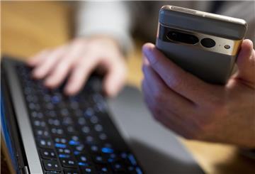 Auf dem Foto ist eine Hand zu sehen, die ein Smartphone hält, die andere liegt auf einer Computertastatur.