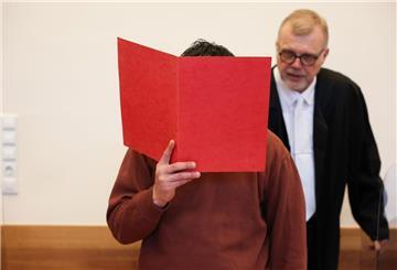 Ein Mann versteckt sein Gesicht hinter einer roten Kladde. Dahinter steht ein Mann mit Anwaltsrobe.