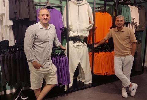 Joschka Traue und Berc Mardiros stehen am Kleiderständer. Auf den Kleiderständern befinden sich Hoodies, T-Shirts und Jogging-Hosen in weiß, lila, orange und grün.