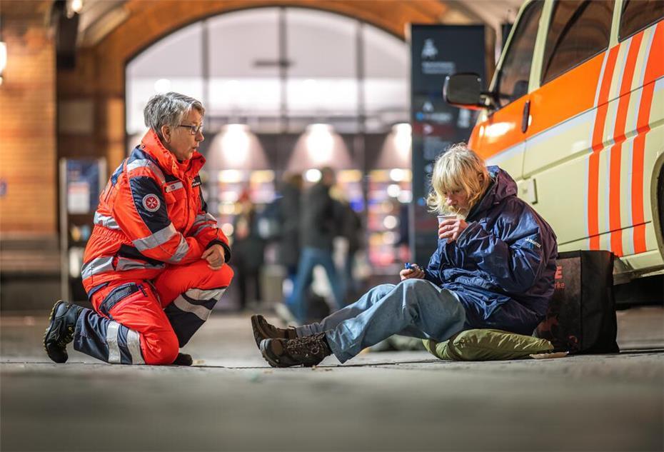Eine Frau von den Johannitern kniet vor einem obdachlosen Mann, der ein Getränk trinkt.