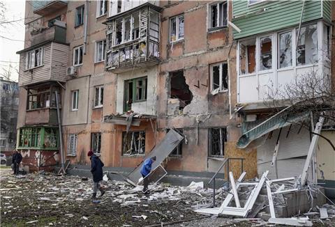 Menschen gehen an einem von Granaten getroffenen Wohnhaus vorüber. 