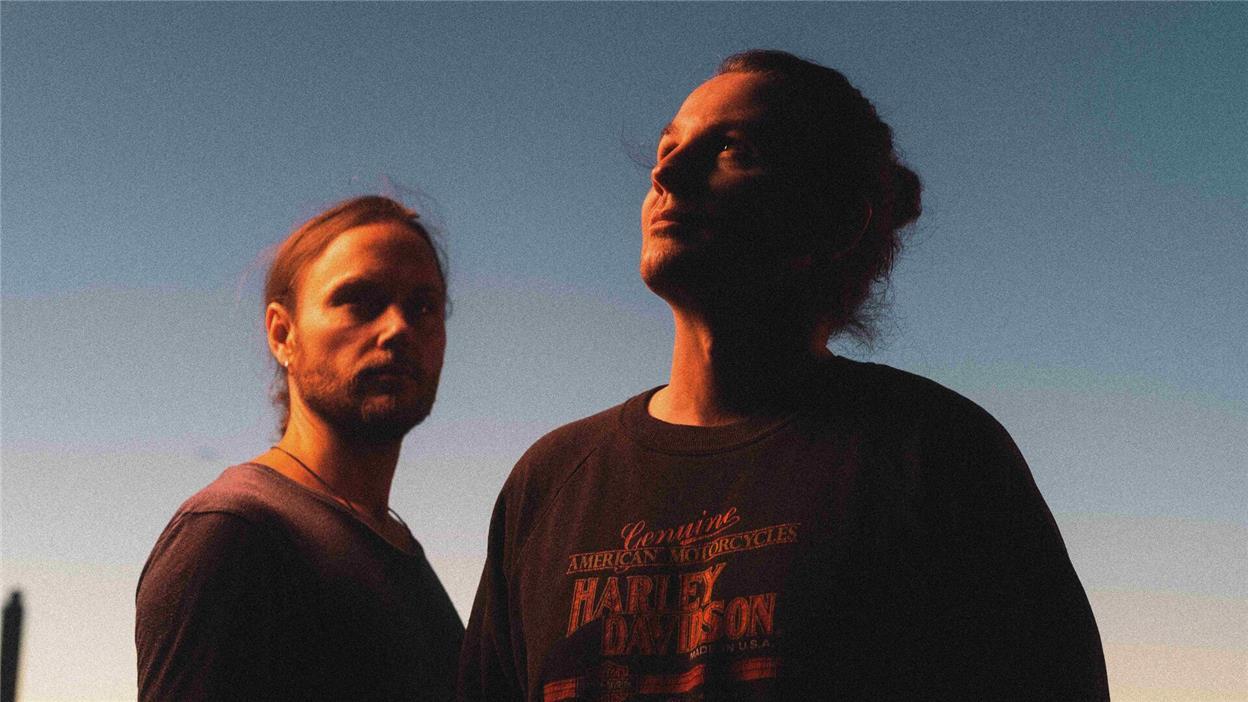 Zwei in Sonnenschein getauchte Männer stehen vor einem blauen Himmel. 