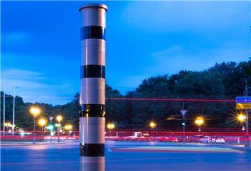 Eine Blitzsäule steht am Kreisverkehr an der Siegessäule, während im Hintergrund vorbeifahrende Fahrzeuge Lichtspuren hinterlassen (Aufnahme mit Langzeitbelichtung).