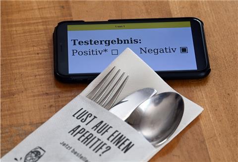 Ein Serviettenetui mit Gabel, Messer und Löffel und dem Aufdruck «Lust auf einen Aperitif?» liegt in einem Restaurant neben einem Smartphone, dessen Display ein negatives Corona-Testergebnis zeigt. 