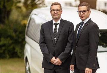 Zwei Männer in schwarzen Anzügen vor weißem Bestattungswagen