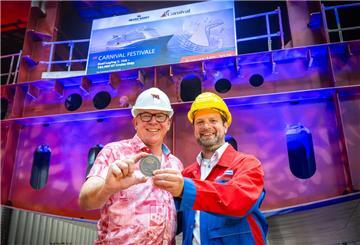 Zwei Männer in Schutzhelmen und Arbeitskleidung halten bei der Kiellegung der „Carnival Festivale“ eine Gedenkmedaille in den Händen. Im Hintergrund ist ein Schiffsrumpf mit einem Bildschirm zu sehen, der den Namen des Kreuzfahrtschiffes anzeigt.