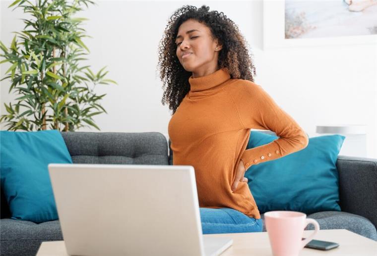Junge Frau mit Rückenschmerzen sitzt vor ihrem Laptop auf dem Sofa.