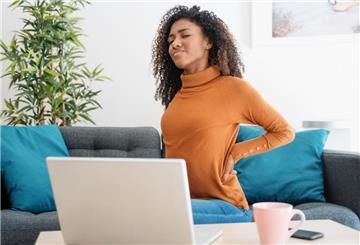 Junge Frau mit Rückenschmerzen sitzt vor ihrem Laptop auf dem Sofa.