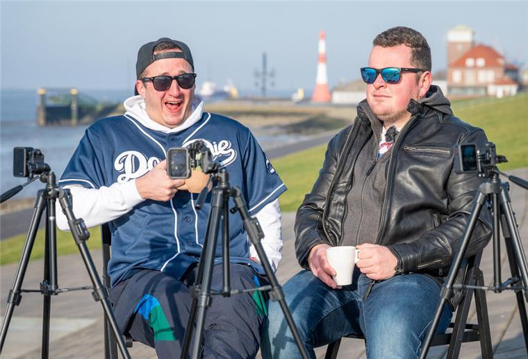 Zwei Männer sitzen auf Stühlen und schauen davor in Kameras. 