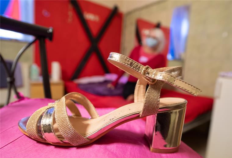 Ein goldener Schuh mit Riemchen und Absatz steht auf einem pinken Stuhlpolster. Dahinter ist schemenhaft eine Frau zu erkennen.