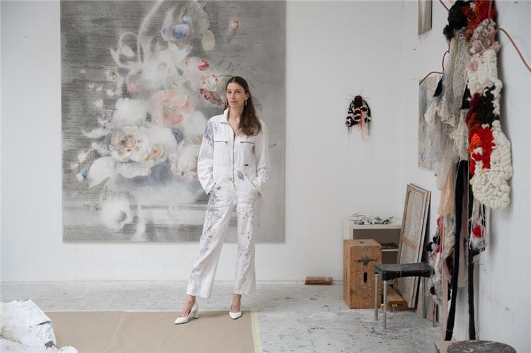 Eine Künstlerin steht vor einem großformatigen Gemälde. Sibylle Springer in ihrem Atelier. An der Wand hinten ist ihrer Interpretation eines Blumen-Stilllebens von Rachel Rysch zu sehen, rechts die Textilarbeit „Harlekinbär“, eigentlich ein kleiner Nachtfalter, der bei Springer riesengroß wird.