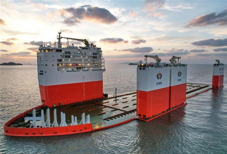 Das halbtauchfähige Schwerlastschiff „Hua Rui Long“ auf See bei Sonnenuntergang, mit geöffnetem Deck und bereit zur Aufnahme eines Großtransports; ruhig liegendes Wasser und Inseln am Horizont.