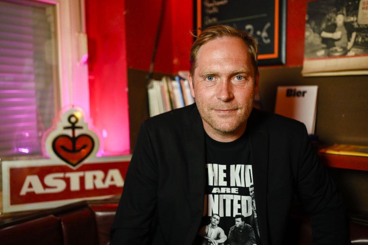 Ein Mann in schwarzem T-Shirt mit weißer Aufschrift und Sakko sitzt vor einem Schild der Biermarke "Astra".