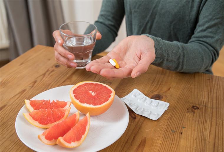 Eine Frau hat Medikamente und ein Glas Wasser in den Händen. Vor ihr steht ein Teller mit Grapefruit.